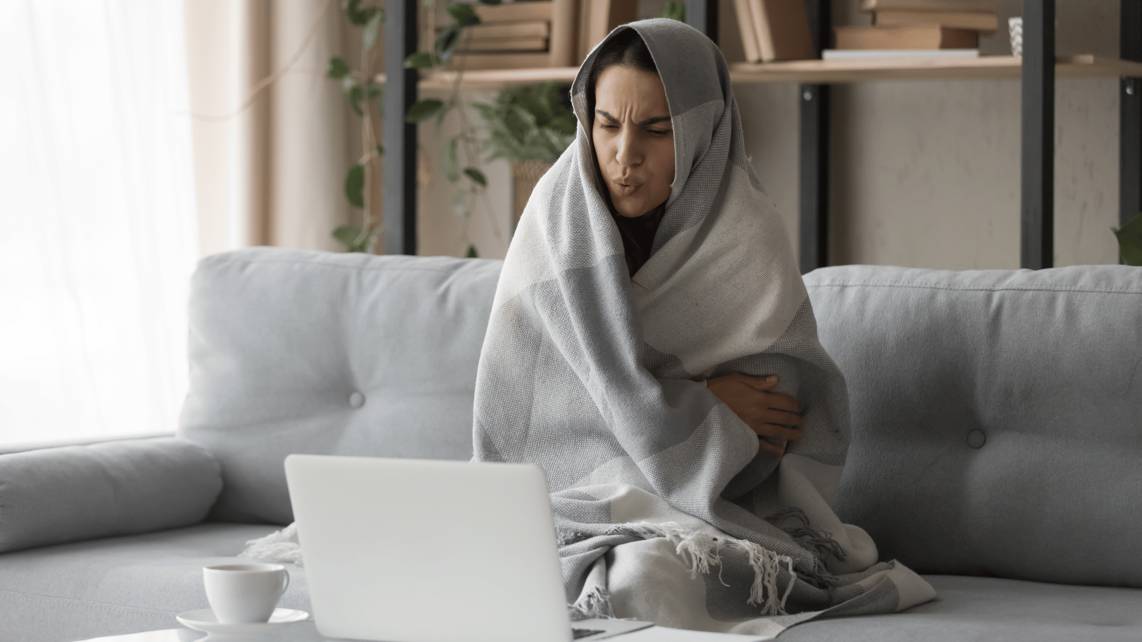 woman sitting in her living room in winter with a blanket over her shoulders and her head searching on her computer for ways to reduce the cost of heating her home.