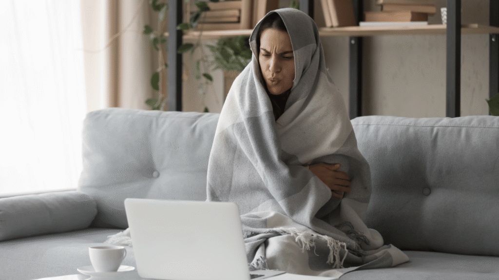 femme assise dans son salon en hiver avec une couverture sur les épaules et la tête qui cherche sur son ordinateur comment réduire le coût de chauffage de sa maison